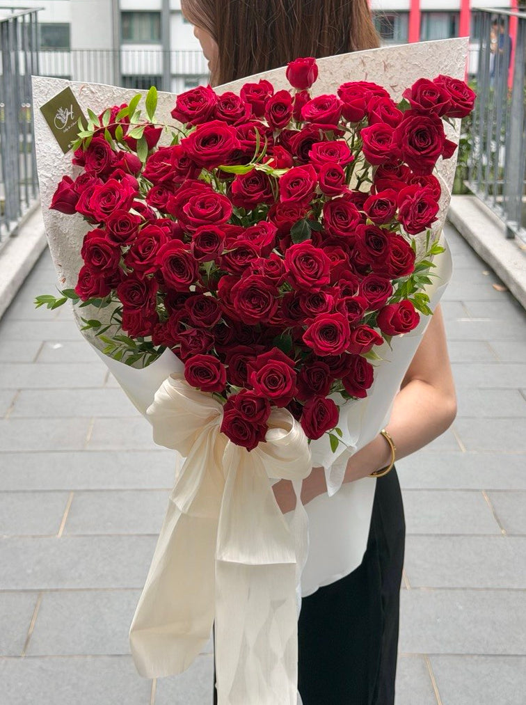A person standing outdoors on a paved walkway holding a large bouquet of deep red roses rose spray wrapped in white paper with a long white ribbon.