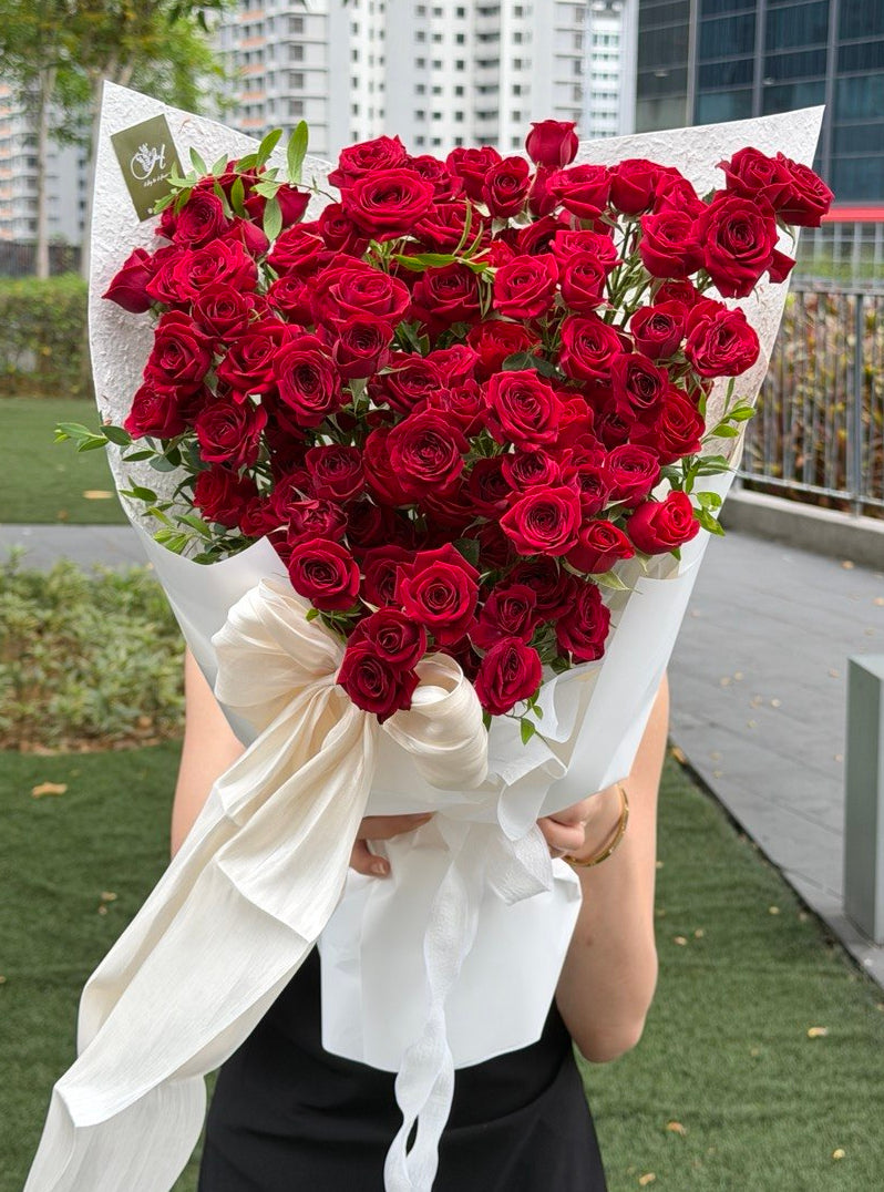 A large heart-shaped bouquet of deep red roses rose spray wrapped in white paper with a white ribbon bow, held by a person standing outdoors on a paved walkway with trees and modern buildings in the background.