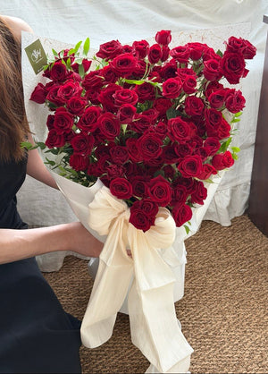 A large bouquet of deep red rose rose spray wrapped in white paper with a cream ribbon bow, held by a person wearing a black dress in an indoor setting with a white draped backdrop and brown carpet.