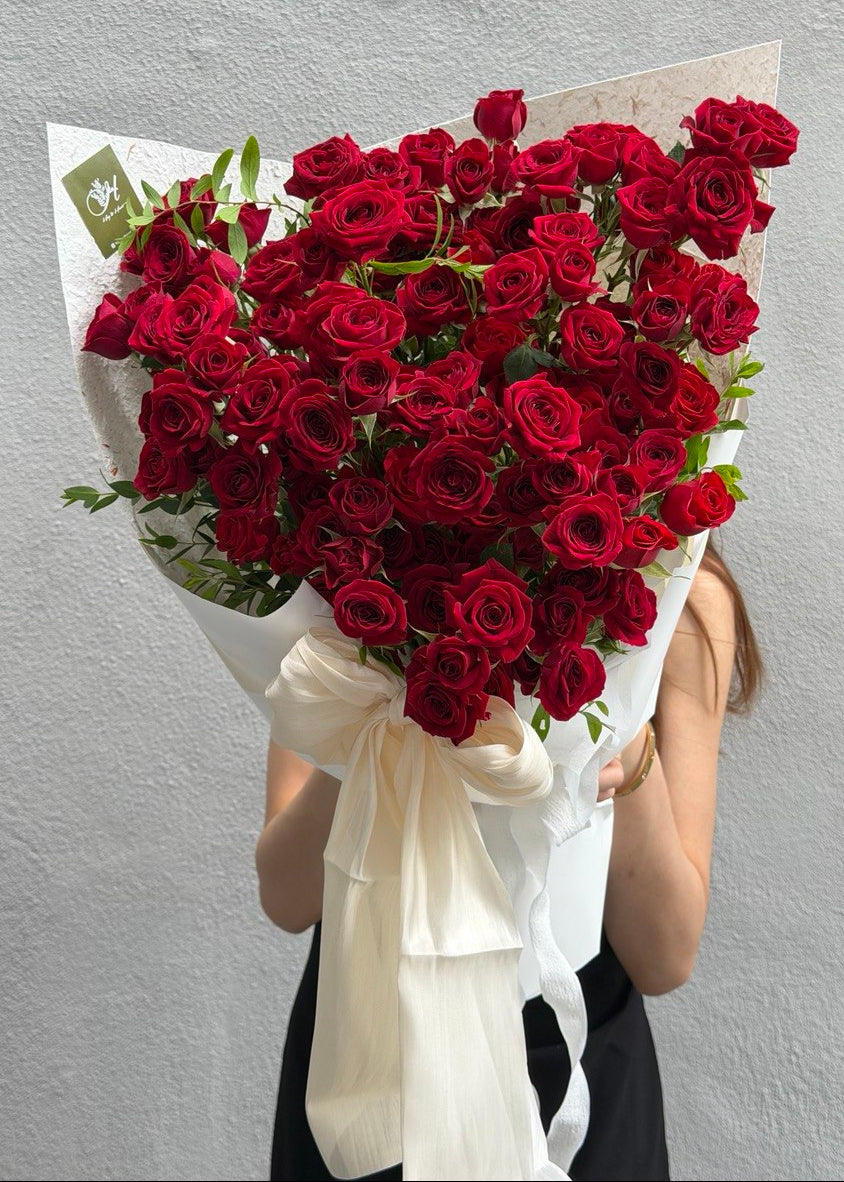 A large bouquet of deep red rose spray wrapped in white paper with a cream ribbon bow, held in front of a person against a light gray textured wall.