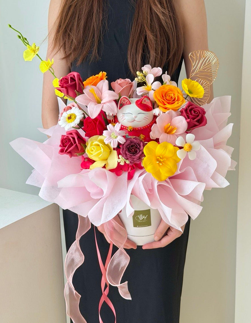A person in a black sleeveless dress holds a large, vibrant artificial flower arrangement in a white pot wrapped with light pink wrapper paper and pink ribbons. The bouquet features a mix of colorful flowers including yellow, orange, red, and white, along with a small white lucky cat figurine and a gold feather accent.