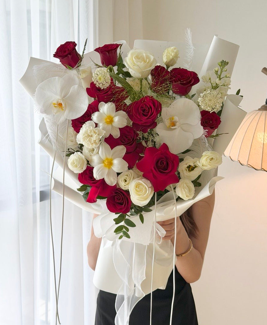 A large, beautifully arranged bouquet of red and white roses, white orchids, and small white flowers wrapped in white paper, held by a person in a black top in a brightly lit room.