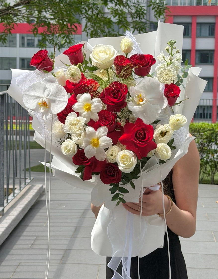 A large bouquet of red and white roses, white orchids, and small white flowers wrapped in white paper, held by a person standing outdoors on a paved walkway with buildings and trees in the background.
