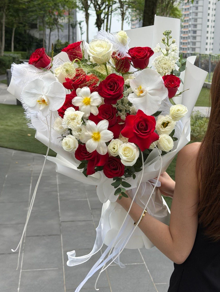 A large bouquet of red and white roses, white orchids, and daisies wrapped in white paper, held by a woman outdoors in front of trees and a building.