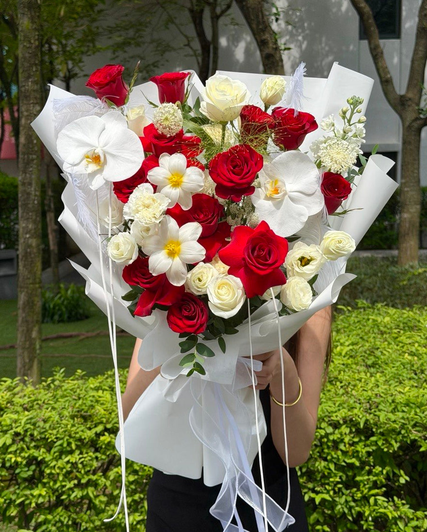 A large floral bouquet wrapped in white paper, held in front of a person's face in an outdoor garden setting. The bouquet features red and white roses, white orchids, and white daisy-like flowers with yellow centers.