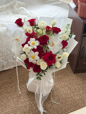 A bouquet of red and white roses, white orchids, and small white flowers wrapped in white tissue paper, placed on a brown carpeted floor next to a sofa.