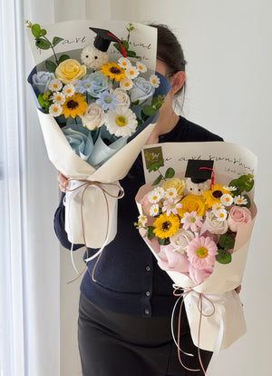 A woman wearing a dark navy cardigan and black skirt holds two graduation-themed flower bouquets wrapped in cream paper. The bouquets feature light blue and yellow roses, white daisies, pink carnations, and cartoon bear figurines wearing graduation caps.