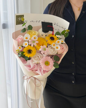 A person in a black button-up shirt holds a wrapped soap flower graduation bouquet featuring yellow sunflowers, pink roses, white daisies, green foliage, and a small plush bear wearing a graduation cap. The bouquet is wrapped in light pink and cream paper with a handwritten card inside.