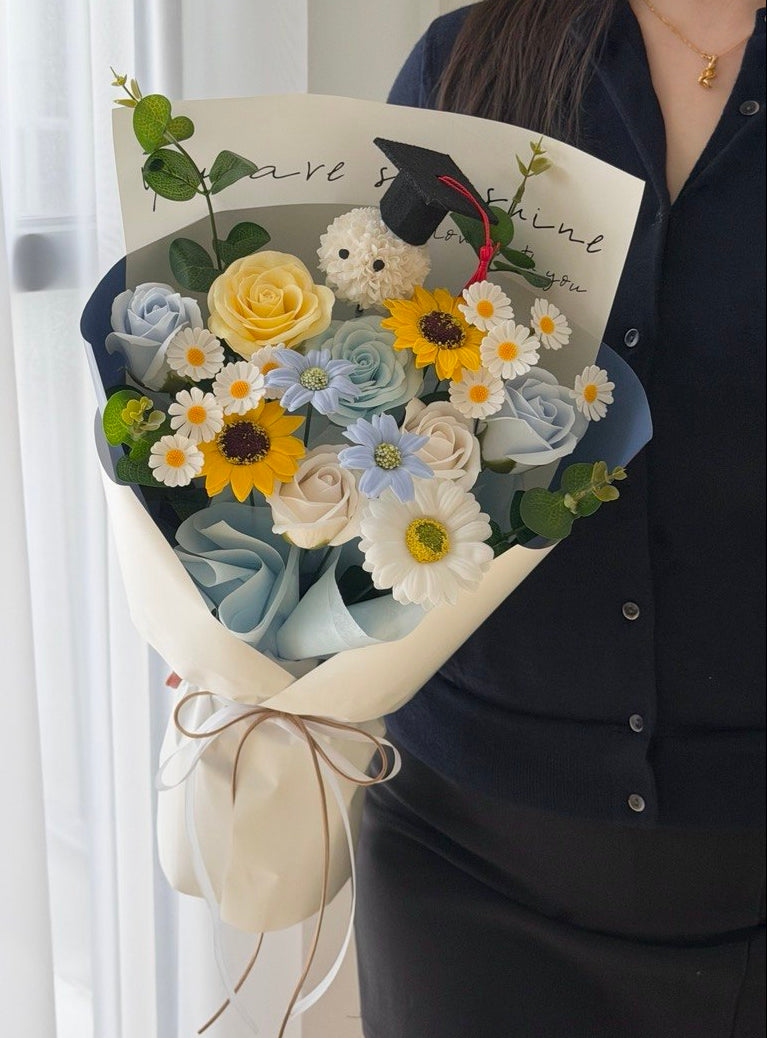 A person in a dark navy shirt holds a beautifully arranged bouquet of soap flowers, including yellow roses, light blue roses, daisies, and a small white plush bear wearing a graduation cap, wrapped in cream paper with a brown twine bow.