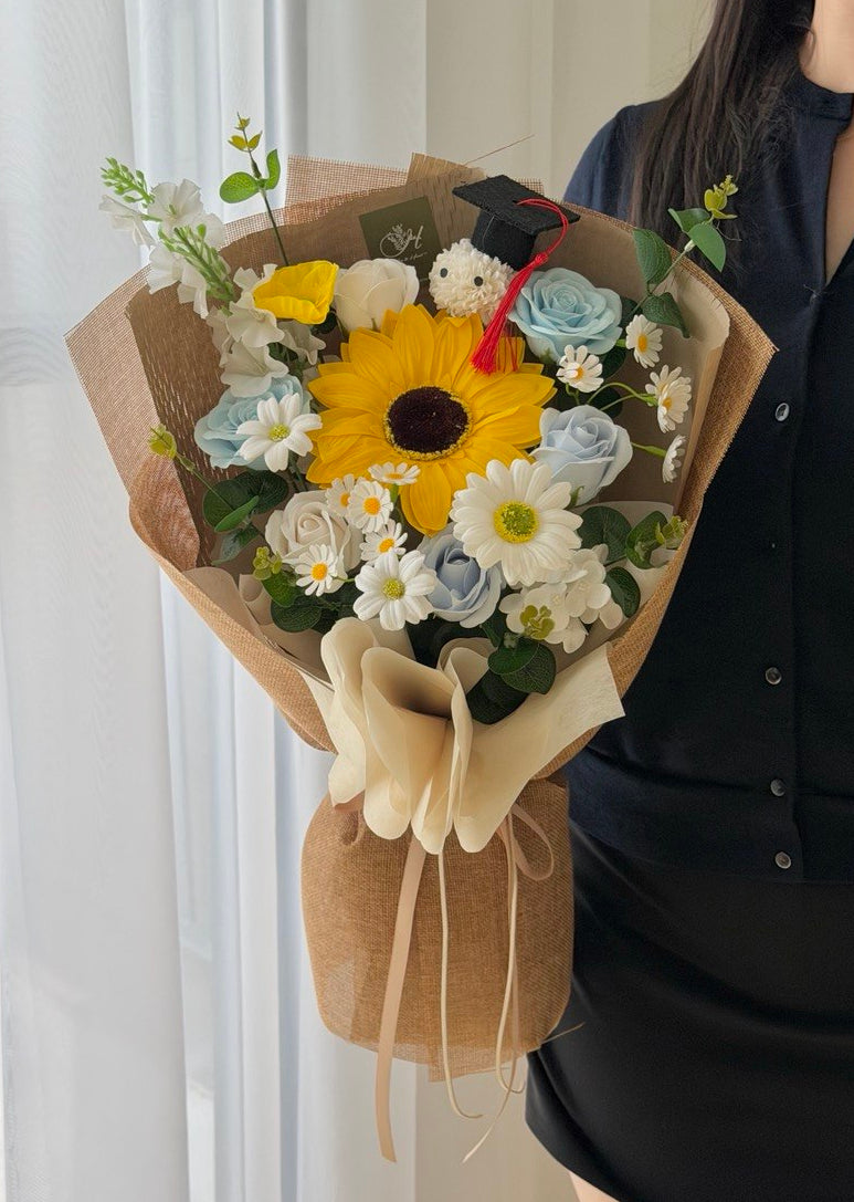 A person in a black shirt holds a wrapped soap flower bouquet featuring a large yellow sunflower, white and light blue daisies, green foliage, and a small graduation cap topper with a red ribbon for graduation flower gifts in KL and Selangor.