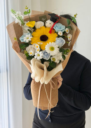 A person in a black long-sleeve top holds a large, beautifully arranged soap flower bouquet wrapped in brown kraft paper. Sunflower graduation bouquet in Sunway University. 