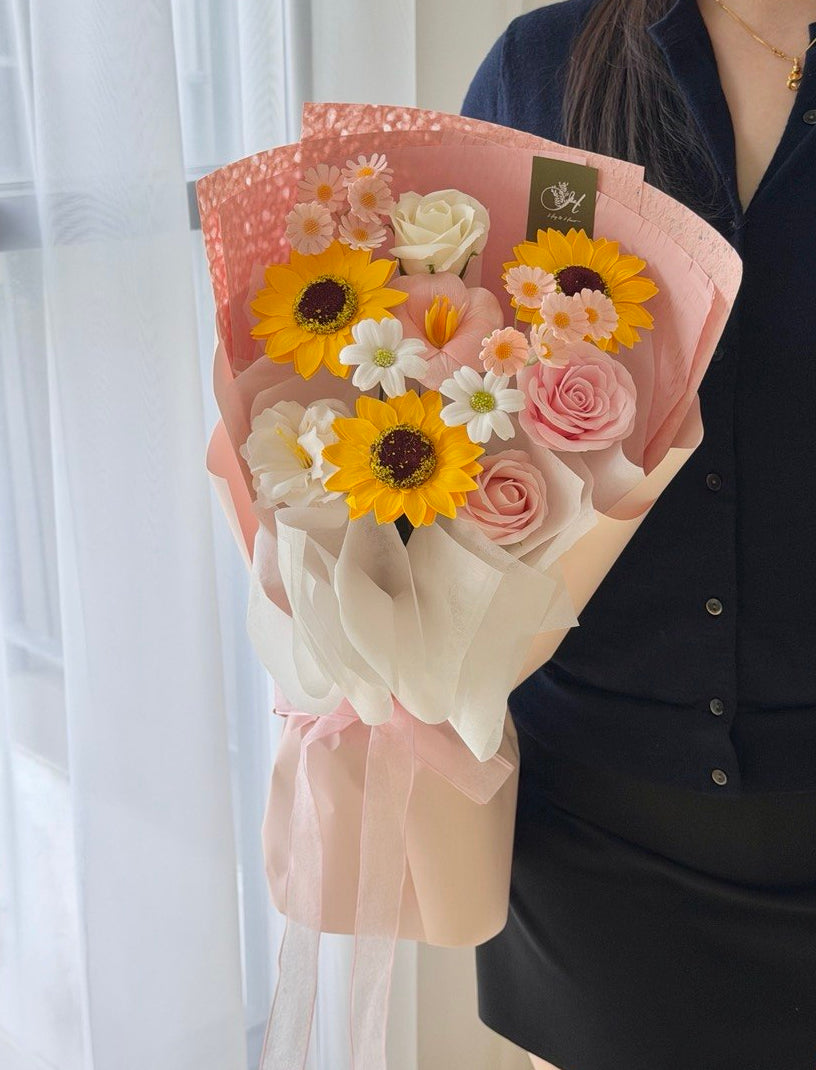 A person in a black shirt holds a wrapped bouquet of flowers featuring yellow sunflowers, pink roses, white daisies, and other small blossoms, wrapped in pink and cream paper, perfect for graduation gift at Subang Jaya.