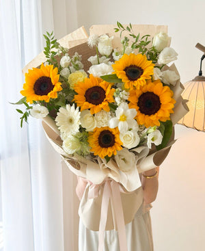 A large floral bouquet featuring bright yellow sunflowers, white roses, white daisies, and green foliage, wrapped in beige paper with a pale pink ribbon, held by a person wearing a light cream dress in a softly lit indoor setting.