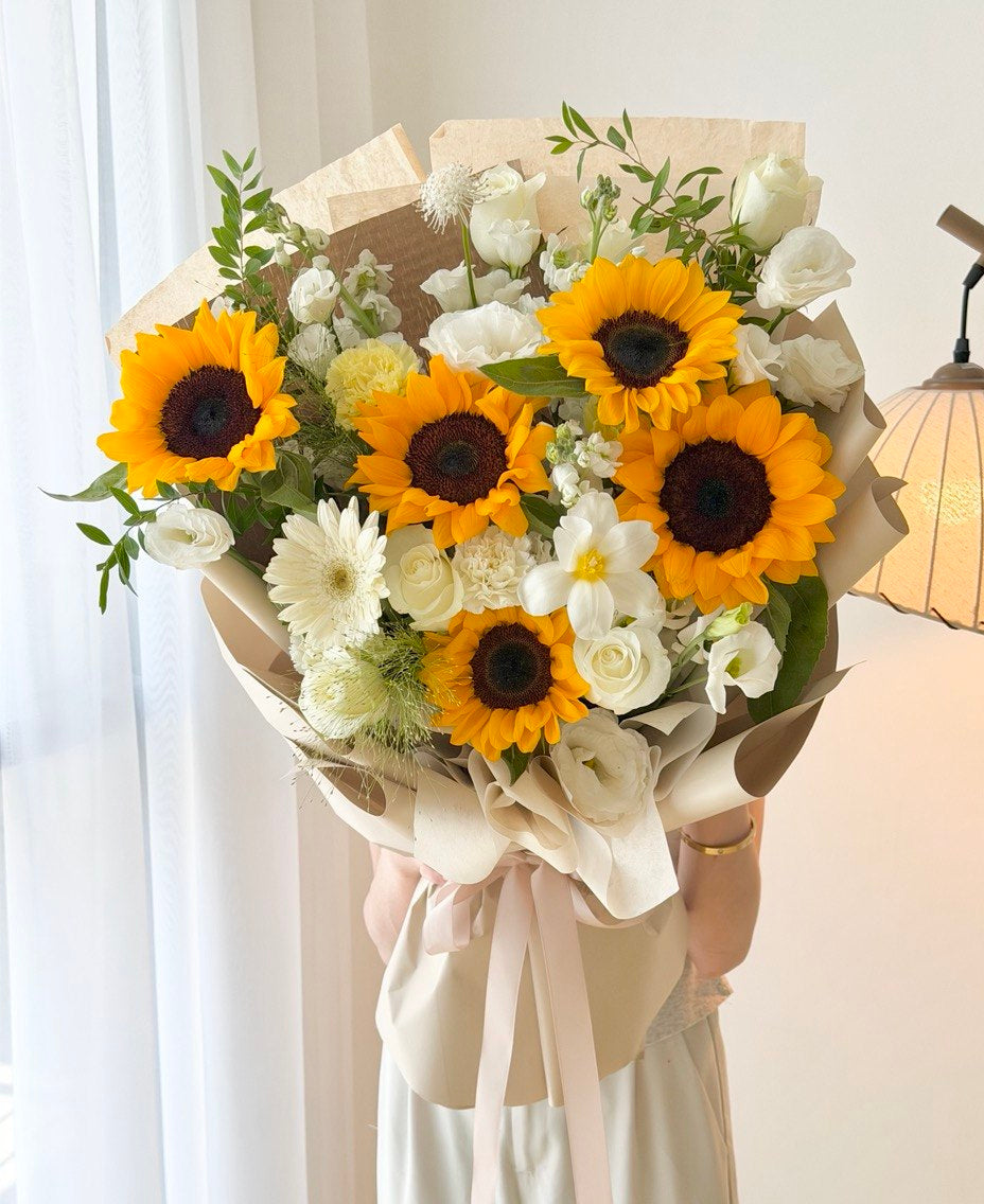 A large floral bouquet featuring bright yellow sunflowers, white roses, white daisies, and green foliage, wrapped in beige paper with a pale pink ribbon, held by a person wearing a light cream dress in a softly lit indoor setting.