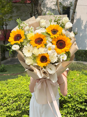 A large floral bouquet held in front of a person's face, featuring bright yellow sunflowers, white roses, and white daisies wrapped in beige paper with a pale pink ribbon. The bouquet is photographed outdoors in front of green hedges and trees.
