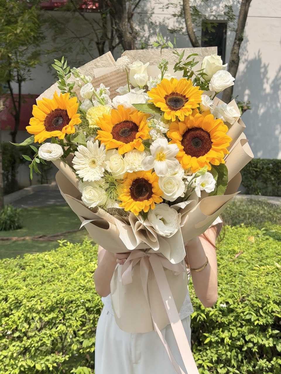 A large floral bouquet held in front of a person's face, featuring bright yellow sunflowers, white roses, and white daisies wrapped in beige paper with a pale pink ribbon. The bouquet is photographed outdoors in front of green hedges and trees.