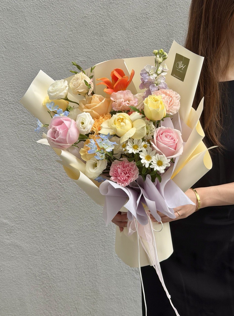 A person holding a large, beautifully arranged floral bouquet with pink, yellow, peach, and white flowers wrapped in cream paper with lavender ribbons, set against a neutral gray wall.