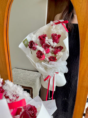 Bouquet of red roses with white accents in a decorative paper wrap, held by a person in front of a mirror.