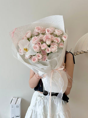 Person holding a bouquet of pink kiss of fairy rose spray wrapped in white paper against a plain background.