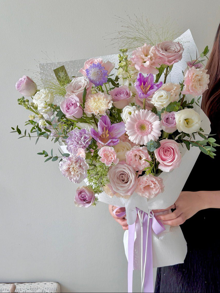 Bouquet of pastel purple theme flowers held by a person against a neutral background.