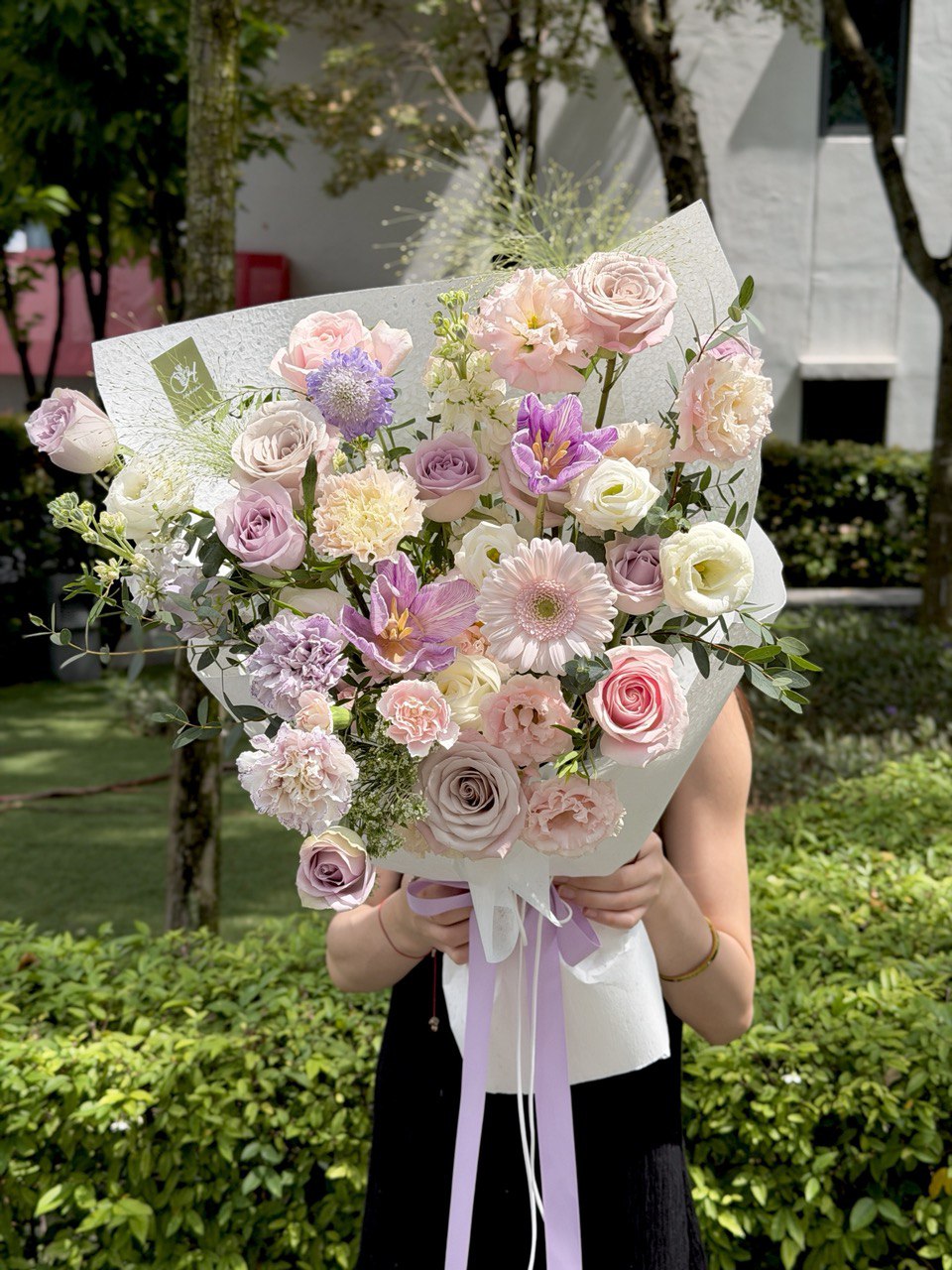 A purple theme flower bouquet with purple tulips, purple carnation, menta roses, lilac roses and some lace flowers is  held by a girl outdoors with greenery in the background.