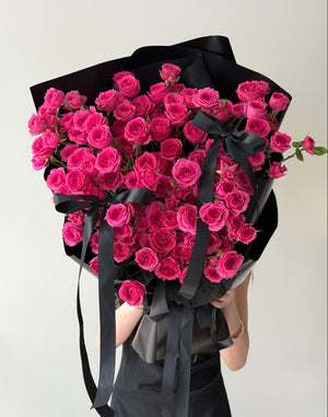 Bouquet of hot pink rose spray wrapped in black paper with a black ribbon held by a person against a white background