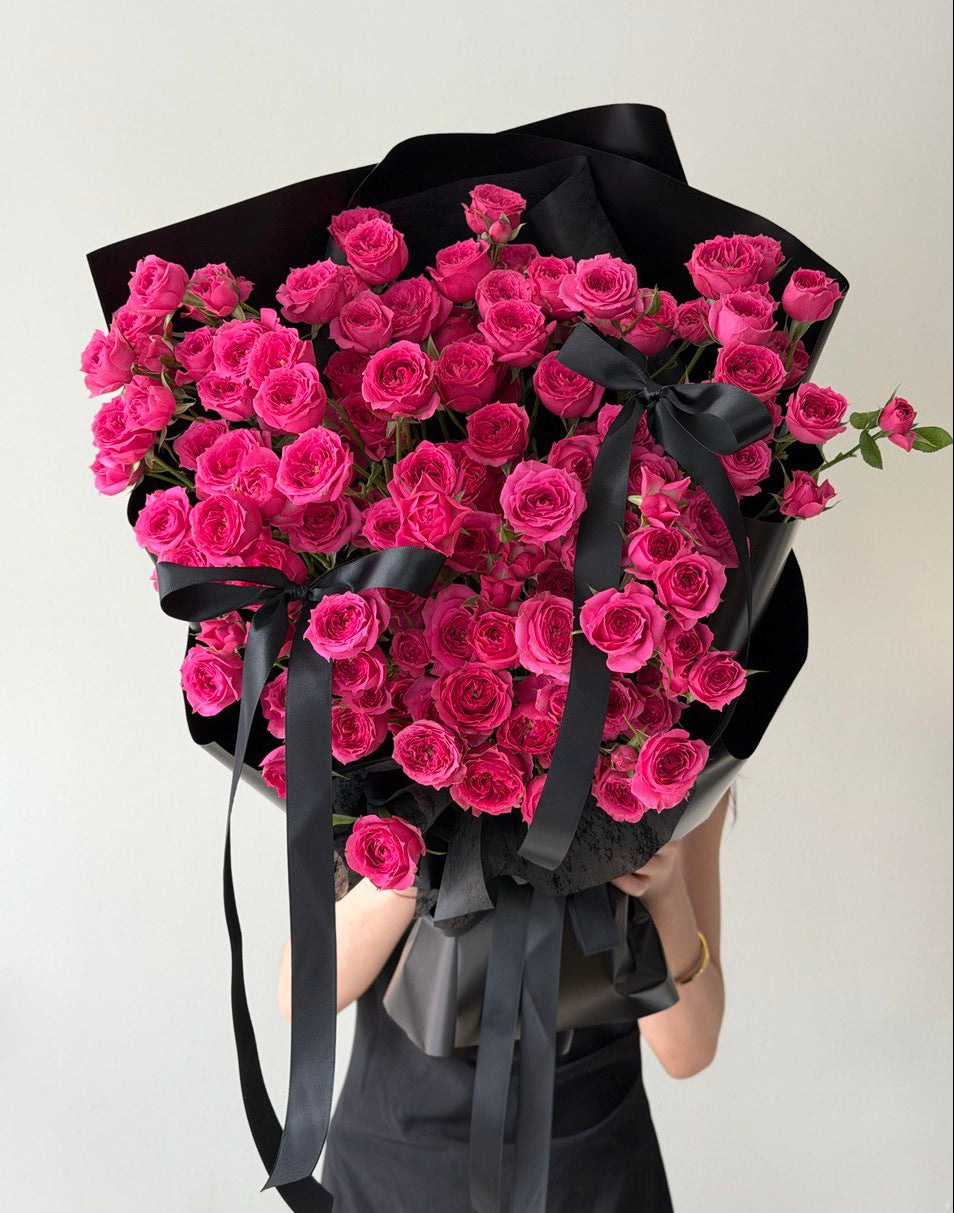 Bouquet of hot pink rose spray wrapped in black paper with a black ribbon held by a person against a white background