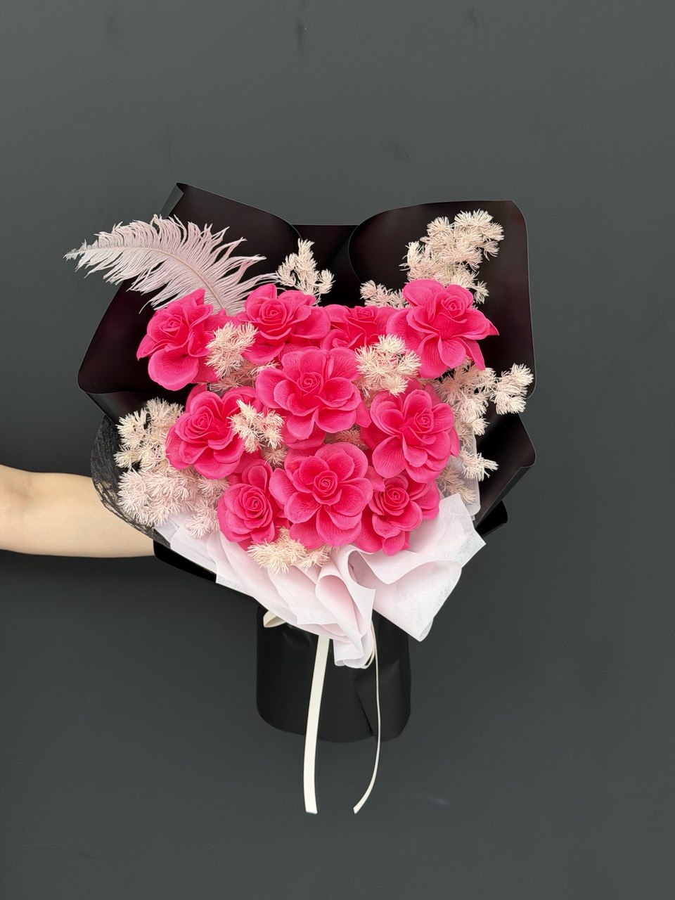 A heart-shaped bouquet of vibrant pink soap roses and dried white filler flowers, wrapped in black and white paper with a feather accent, held by a hand against a dark gray background.