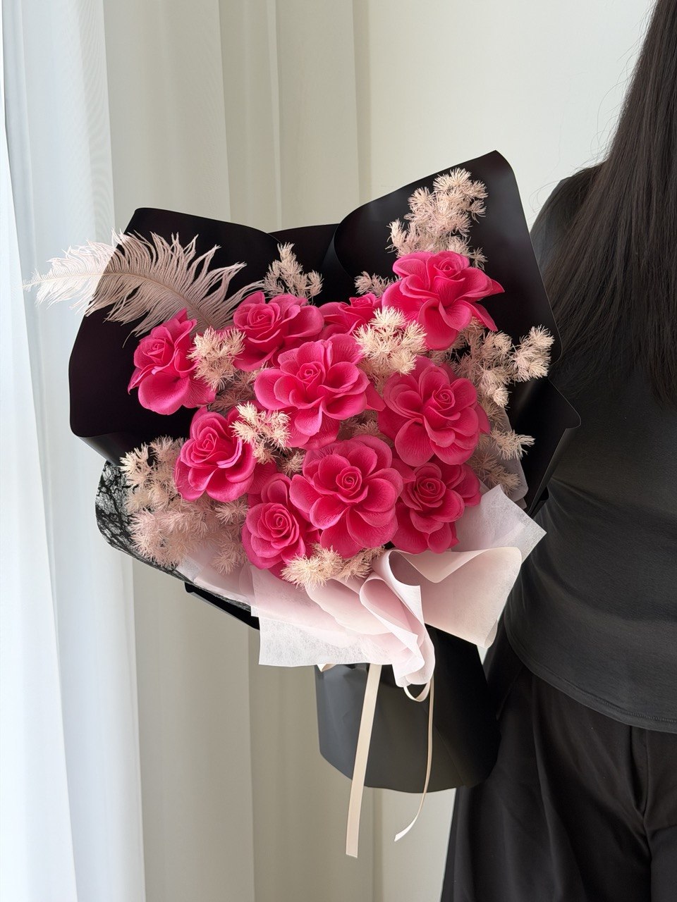 A bouquet of pink rose soap flower wrapped in black paper, held by a person wearing a black top and black skirt in front of a white curtain.