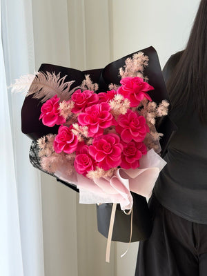 A bouquet of pink rose soap flower wrapped in black paper, held by a person wearing a black top and black skirt in front of a white curtain.