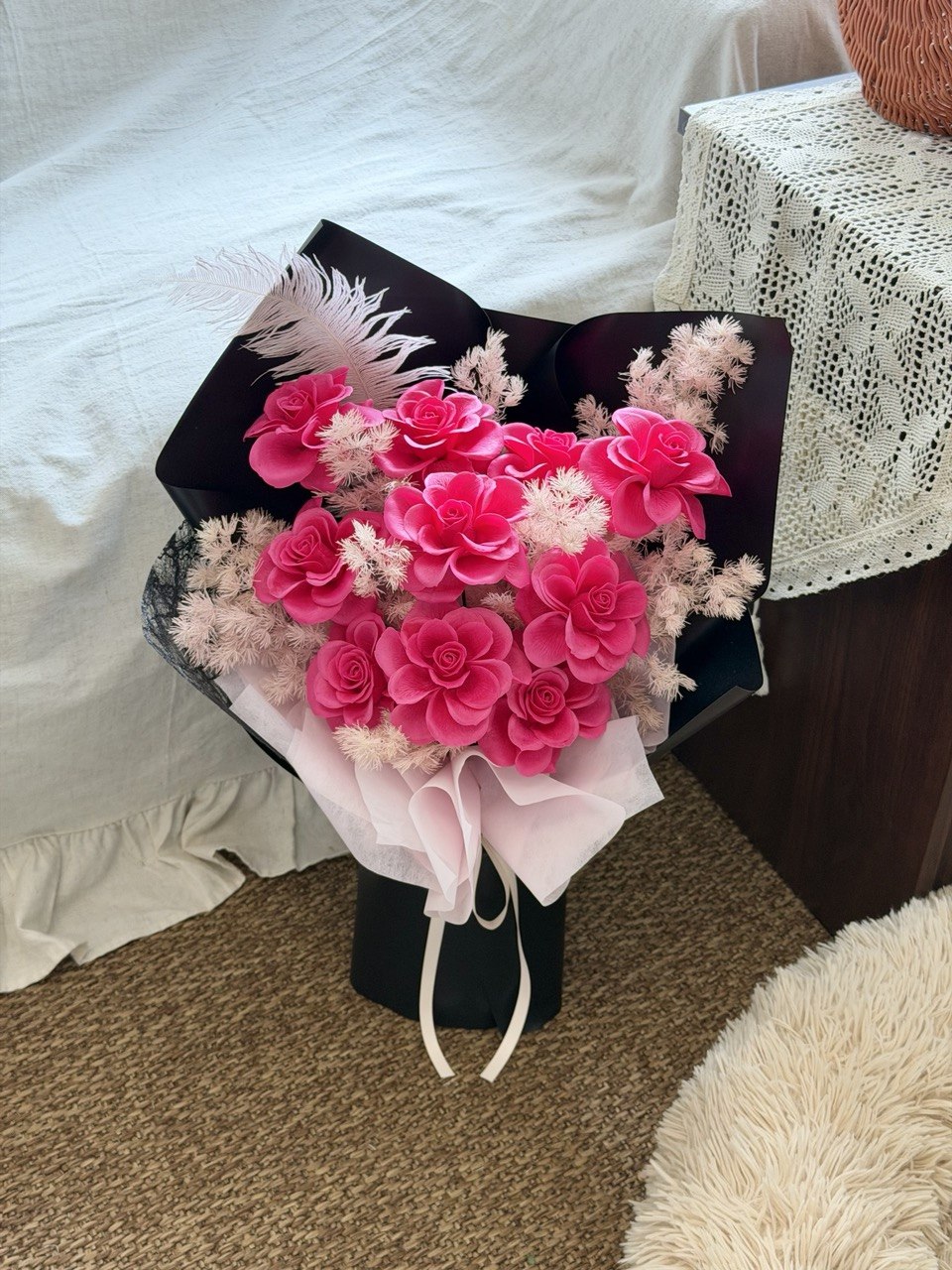 A bouquet of pink soap flowers arranged in a black box with pale pink tissue paper and a white feather, photographed on a carpeted floor beside a bed and a lace-covered table.