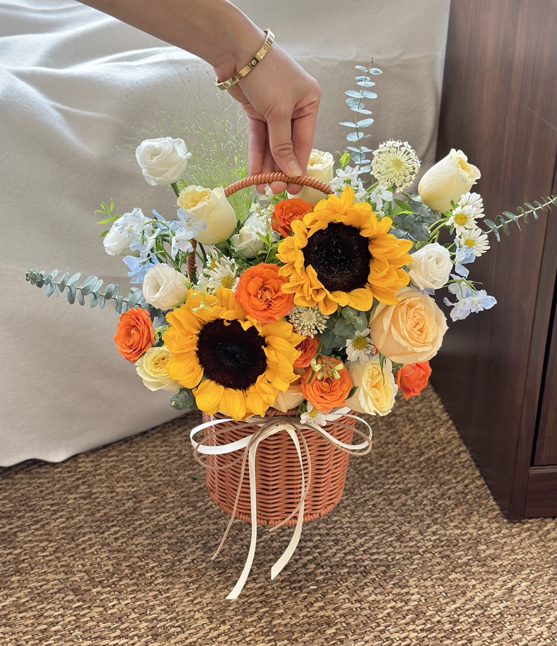 Bouquet of flowers basket consist of sunflower, buttercup rose, champagne rose,  delphinium blue in a basket held by a person on a carpeted floor.