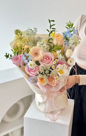 Bouquet of flowers with light pink roses, delicate white chamomile, delphinium blue, and soft peach carnations held by a person in a white room.