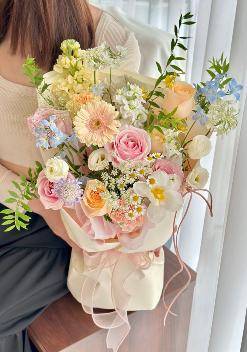 A pastel flower bouquet featuring pink roses, gerbera daisies, champagne rose, peach and cream blooms, wrapped in soft white and pink paper.

