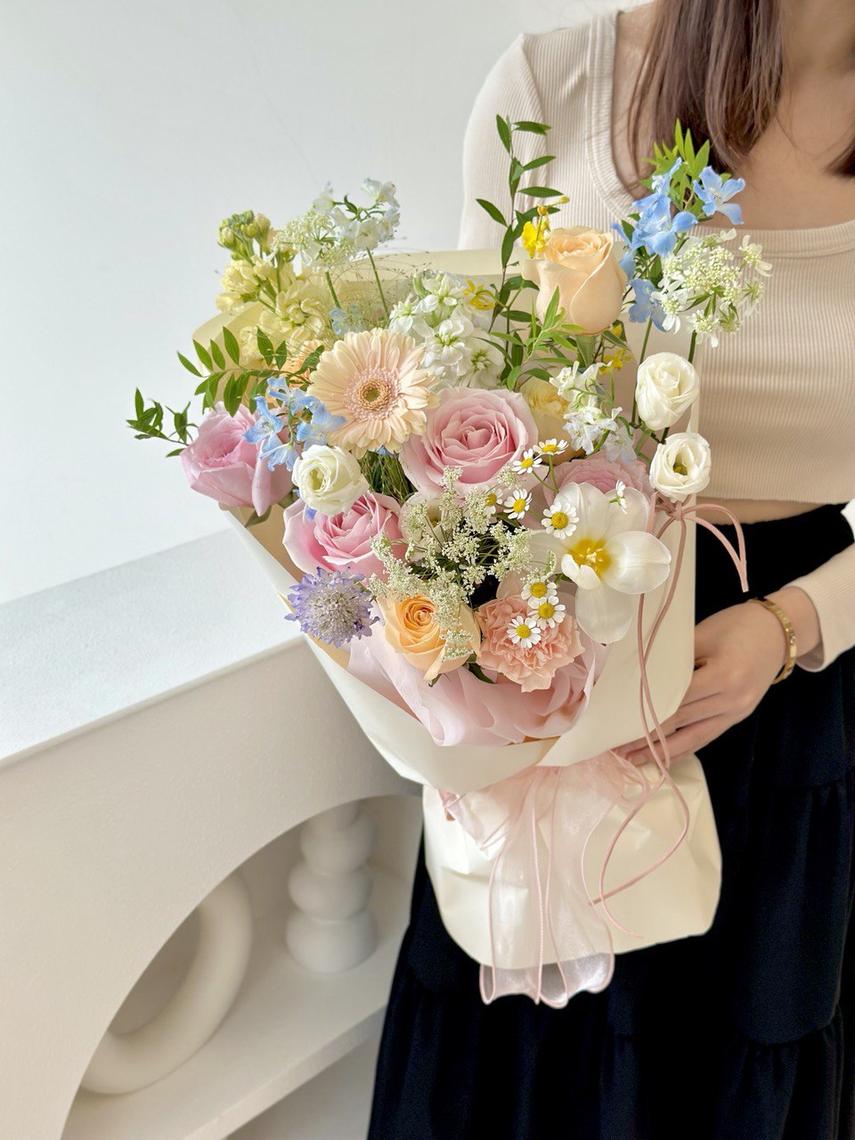 Person holding a bouquet of colorful flowers with shades of blush, ivory, and soft blue blooms tied with a sheer pink ribbon held against a light background.