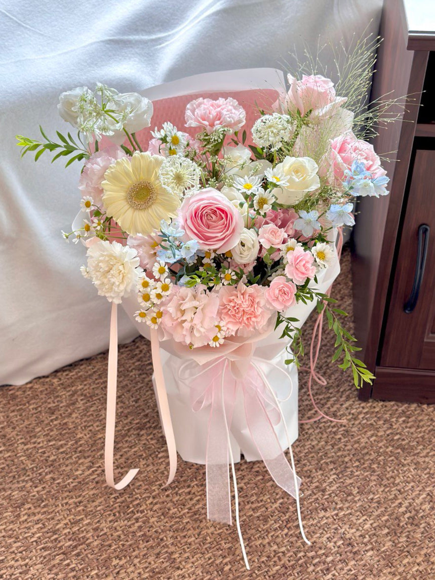 A soft pastel bouquet featuring pink roses, white gerberas, carnations, and chamomile blooms, elegantly wrapped with long pink ribbons.