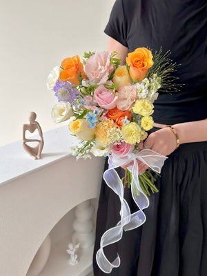 Charming hand-tied bridal bouquet with pastel blooms, orange rose, and fresh greenery, wrapped with ribbon for weddings.
