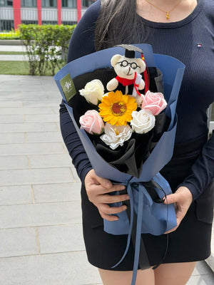 A person outdoors holding a wrapped flower bouquet with a small graduation bear wearing a graduation cap and gown, pink and white roses, a yellow sunflower, and white rose soap flowers. The bouquet is wrapped in blue paper with a black inner layer and ribbon.