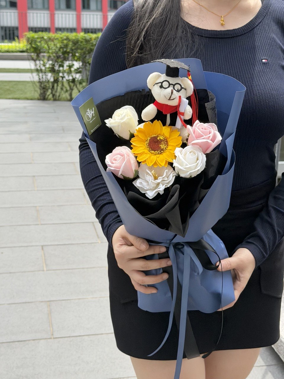 A person outdoors holding a wrapped flower bouquet with a small graduation bear wearing a graduation cap and gown, pink and white roses, a yellow sunflower, and white rose soap flowers. The bouquet is wrapped in blue paper with a black inner layer and ribbon.