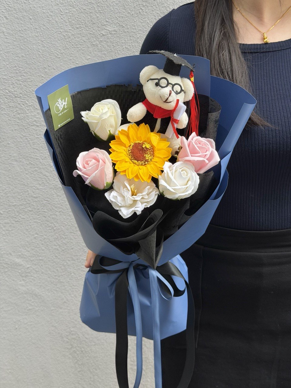A graduation-themed soap flower bouquet wrapped in blue and black paper, featuring a yellow sunflower, pink and white roses, a small plush graduation bear in a graduation cap, and a blue ribbon bow. A person wearing a dark blue ribbed top is holding the bouquet against a light gray wall.