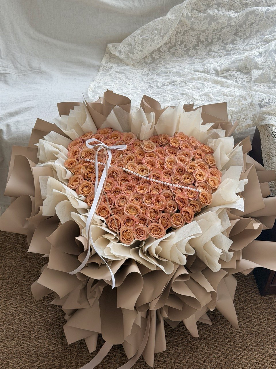 A heart-shaped bouquet of cappuccino rose wrapped in beige and khaki paper, tied with a white ribbon, placed on a carpeted floor in front of a white bed.
