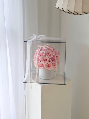A round arrangement of light pink roses in a white box, enclosed in a clear square gift box with a white ribbon, displayed on a white pedestal in a bright indoor setting.