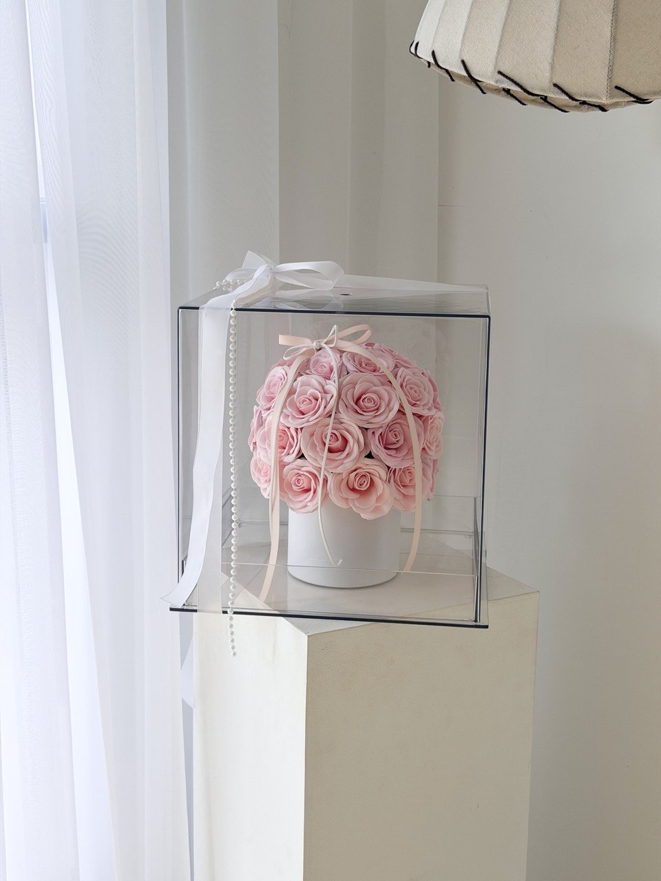 A round arrangement of light pink roses in a white box, enclosed in a clear square gift box with a white ribbon, displayed on a white pedestal in a bright indoor setting.
