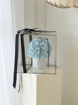 A clear square gift box containing a round arrangement of light blue artificial roses on a white box, tied with a long black ribbon and a pearl bead chain, displayed on a white pedestal.
