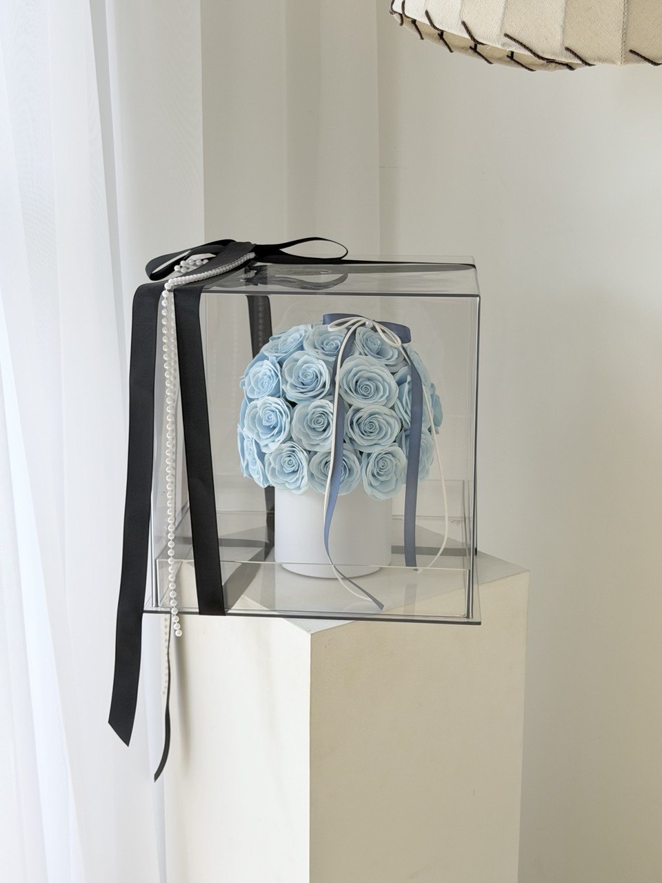 A clear square gift box containing a round arrangement of light blue artificial roses on a white box, tied with a long black ribbon and a pearl bead chain, displayed on a white pedestal.