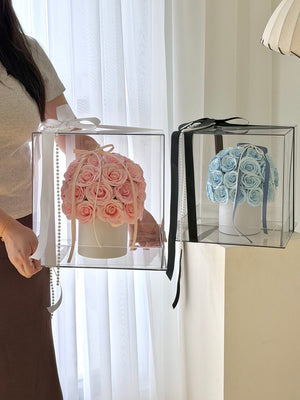 Two clear acrylic flower boxes with black and white ribbons are shown indoors. One box contains pink roses, and the other contains light blue roses.