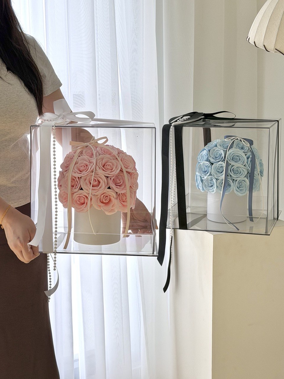 Two clear acrylic flower boxes with black and white ribbons are shown indoors. One box contains pink roses, and the other contains light blue roses.