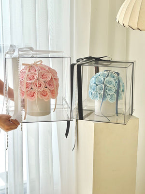 Two clear acrylic flower boxes with rounded corners, one holding pink roses and the other holding light blue roses, shown in a bright indoor setting with white curtains and a lamp.