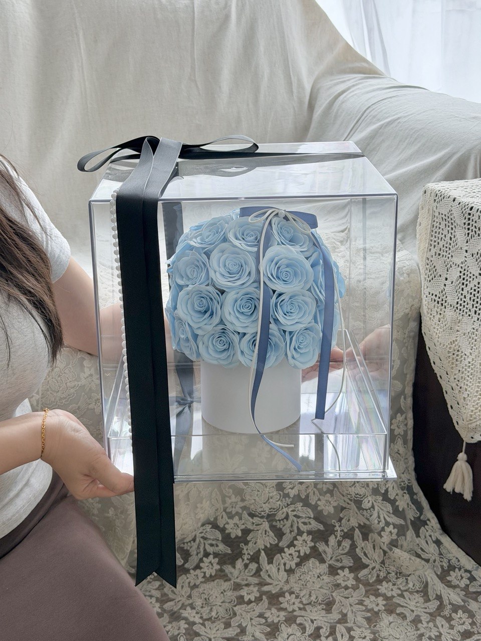 A clear acrylic gift box containing a bouquet of light blue artificial roses on a white vase, tied with a dark blue ribbon, held by a person in a home setting.