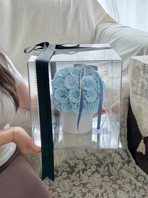 A clear acrylic gift box containing a bouquet of light blue artificial roses on a white vase, tied with a dark blue ribbon, held by a person in a home setting.